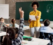 Happy African American teacher holding a class and pointing at student with raised arm who wants to answer the question.